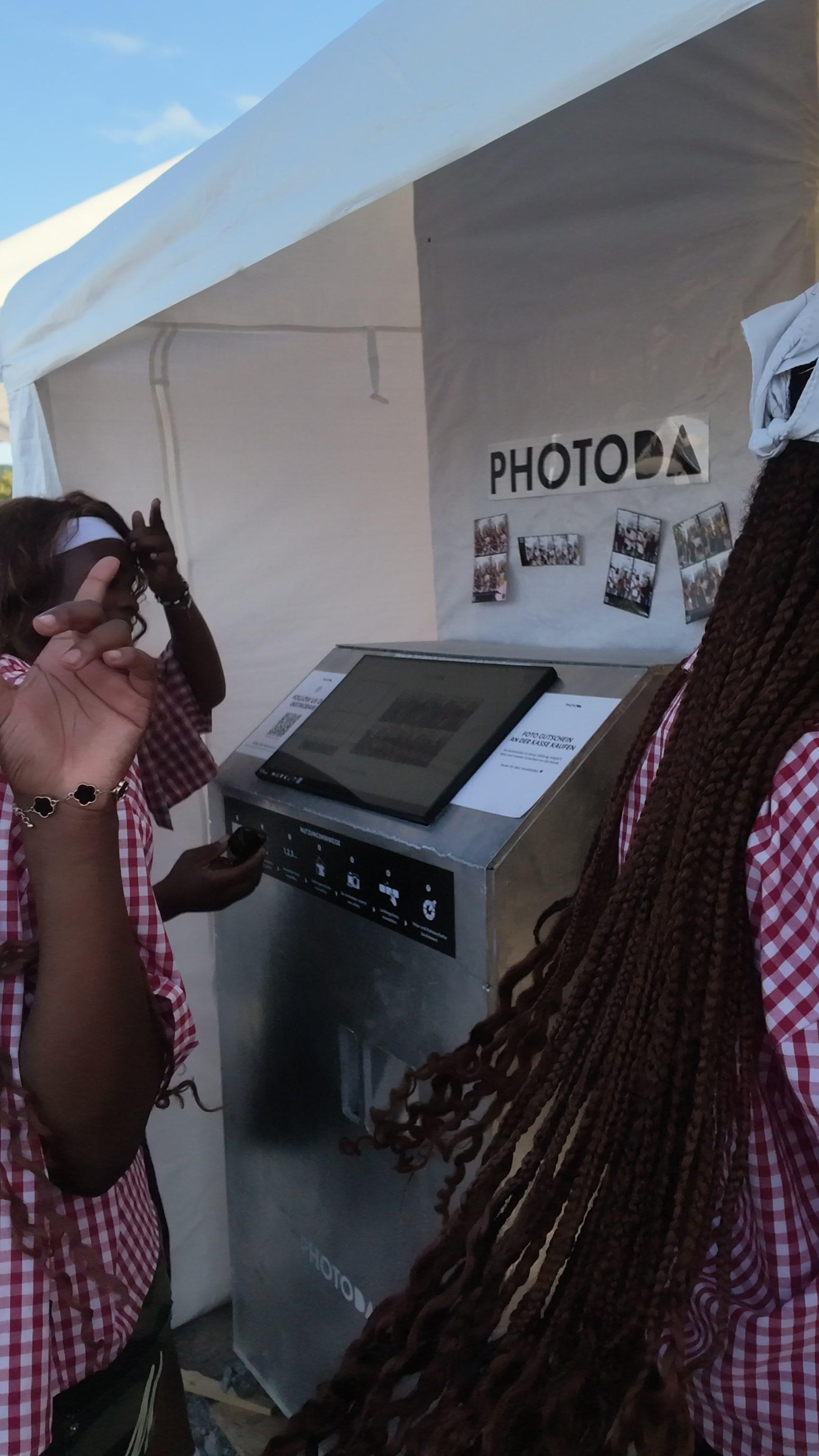 The PhotoDA photo booth kiosk on site at a festival, with on-screen instructions and printed sample strips displayed beside it