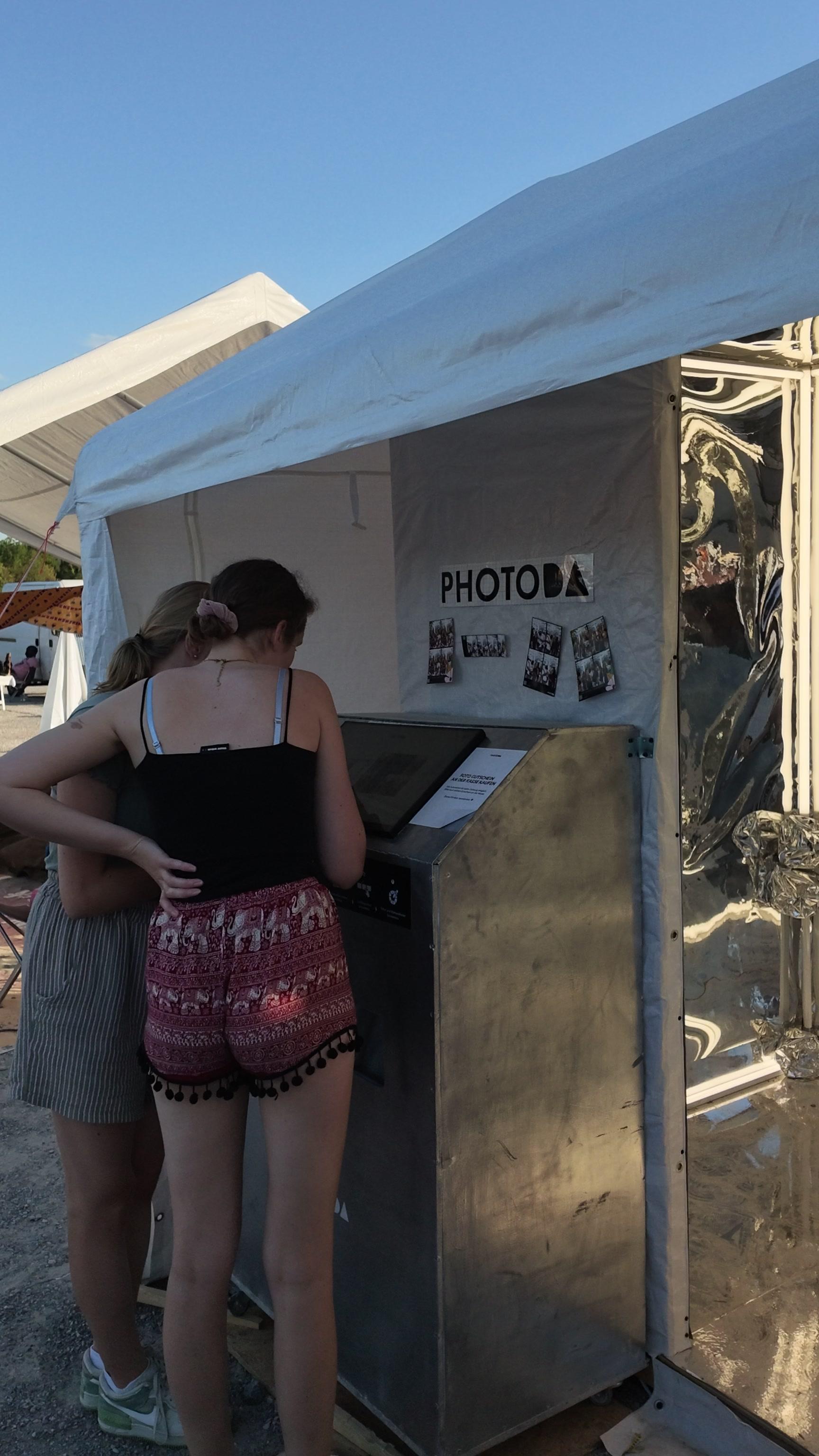 A real PhotoDA photo booth set up at an outdoor event, with branded tent, mirrored entrance and printed photo strips on display